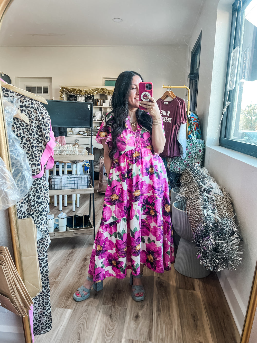 Woman in a pink floral dress taking a mirror selfie in a room with clothing and decor.