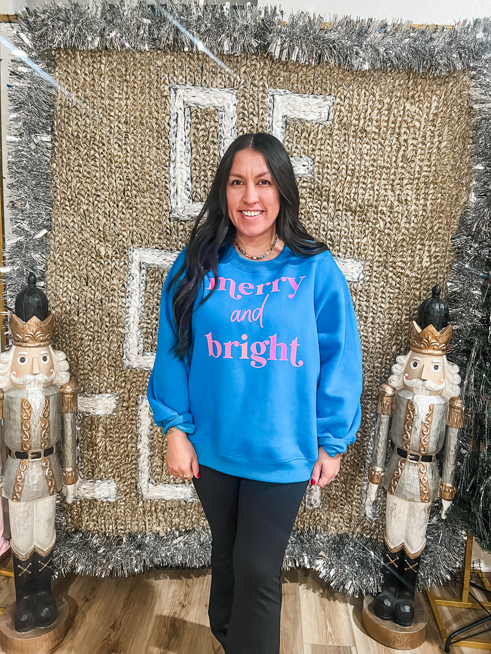 Woman wearing a blue sweatshirt with 'merry and bright' text, standing in front of a decorative background with Nutcrackers.