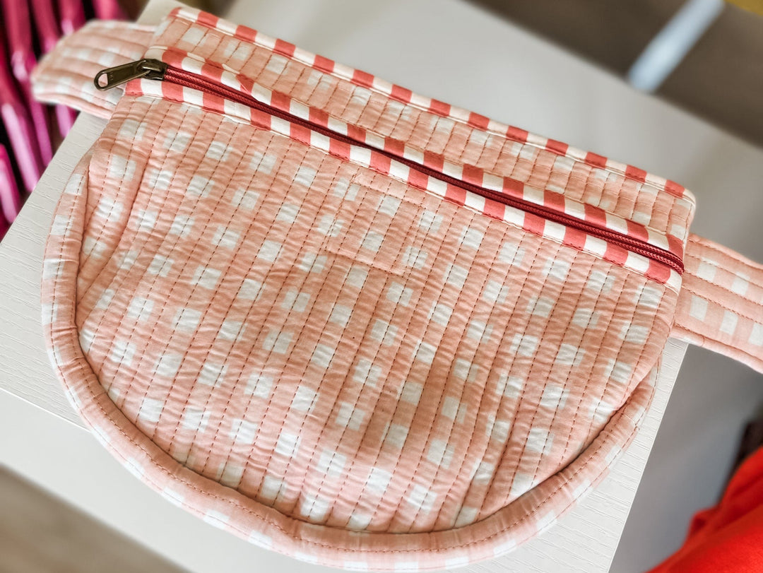 A quilted fabric zippered pouch in a pink and white gingham pattern, shown resting on a white surface in a styled indoor setting.
