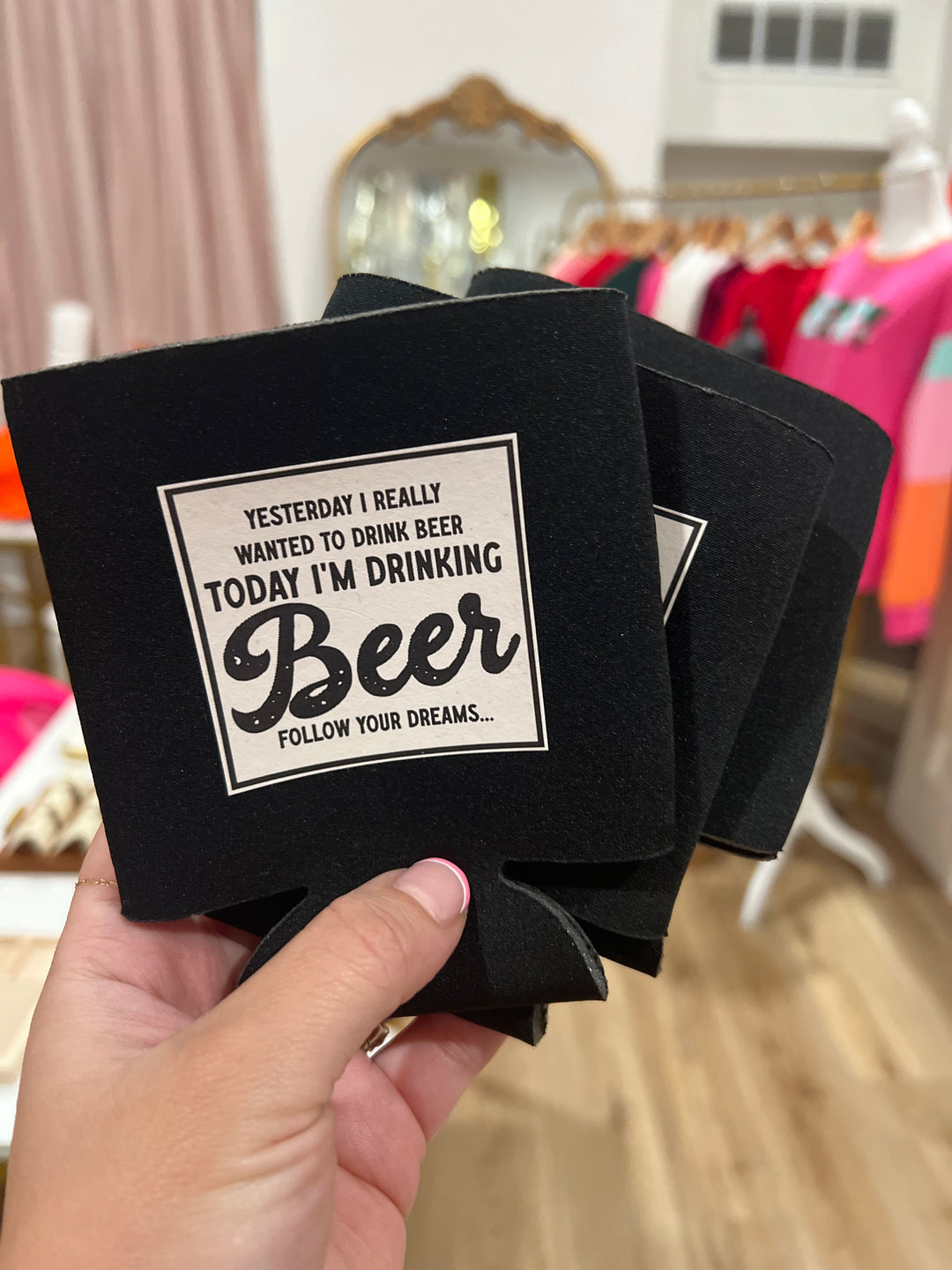 Two black koozies with white text holding a beverage, one koozie shows the front side with the text &