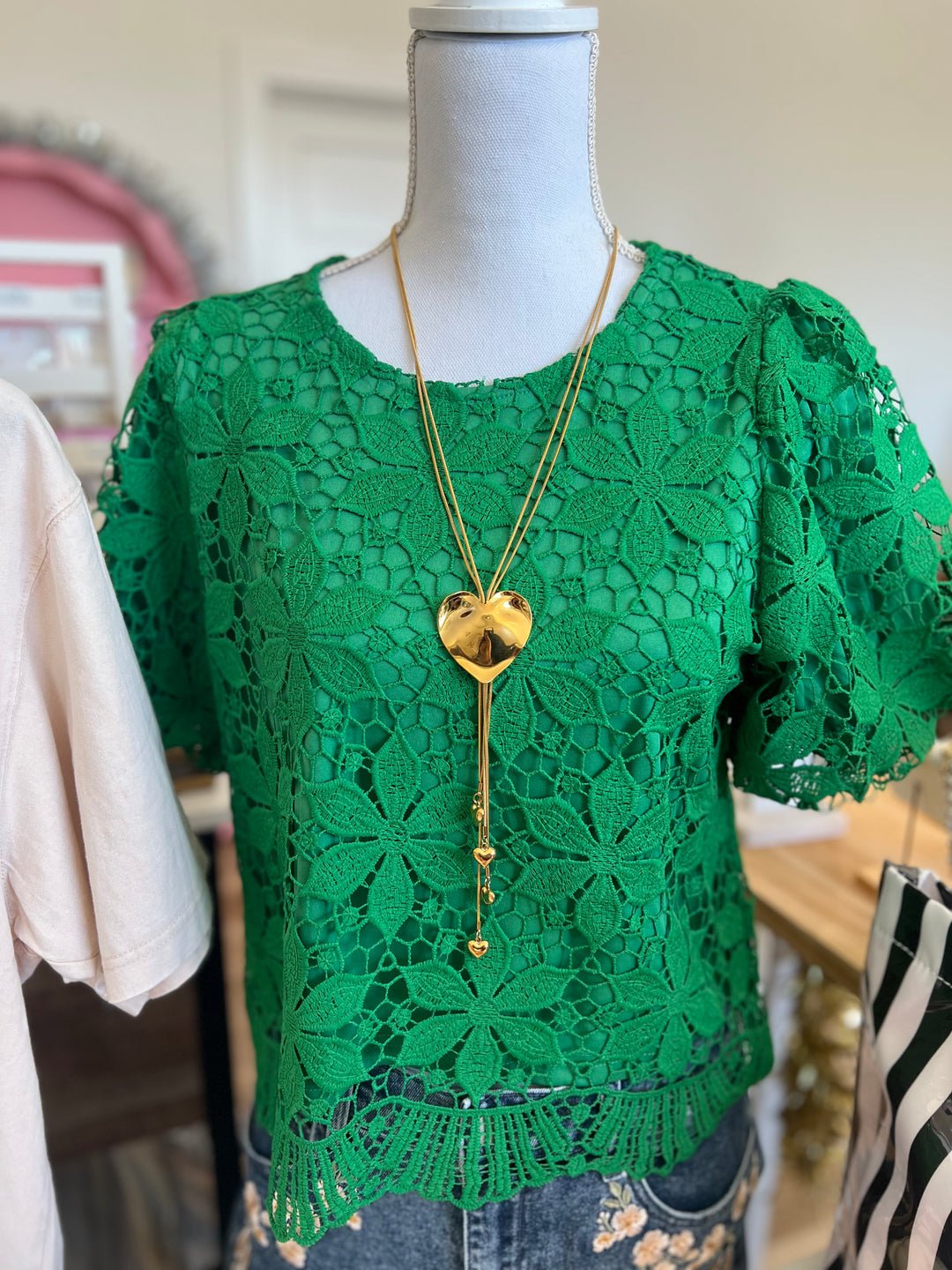 A bright green lace short-sleeve top displayed on a mannequin, styled with a long gold heart pendant necklace and blue jeans.