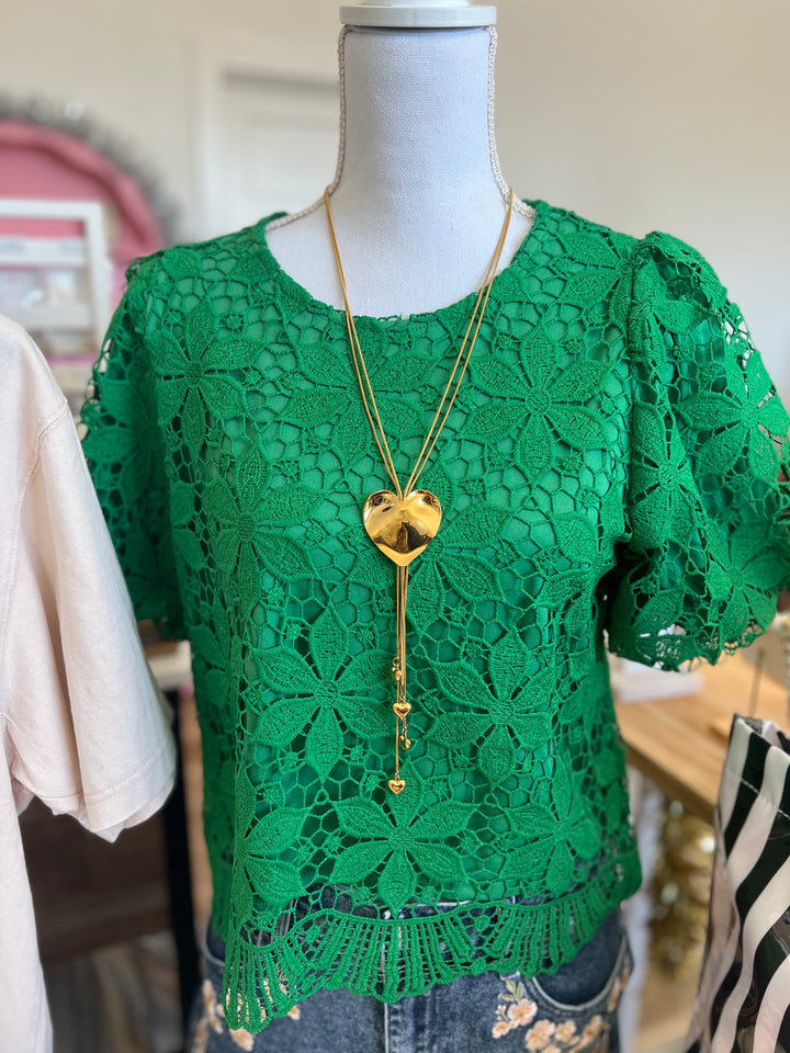 A bright green lace short-sleeve top displayed on a mannequin, styled with a long gold heart pendant necklace and blue jeans.