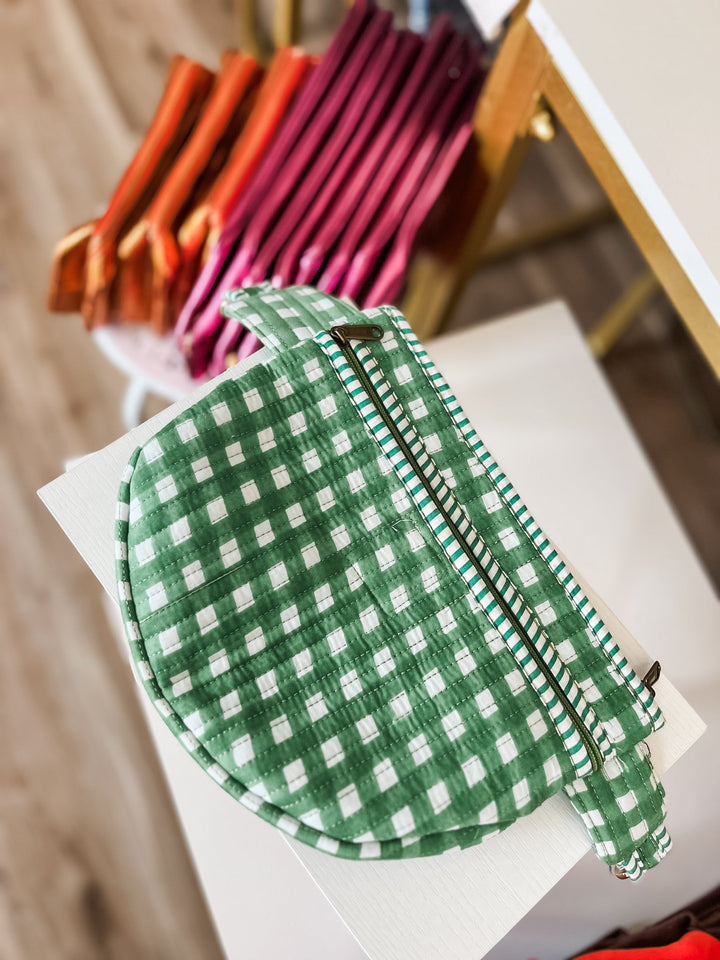A green and white gingham zippered pouch with a matching strap, shown in a styled setting with other colorful pouches blurred in the background.