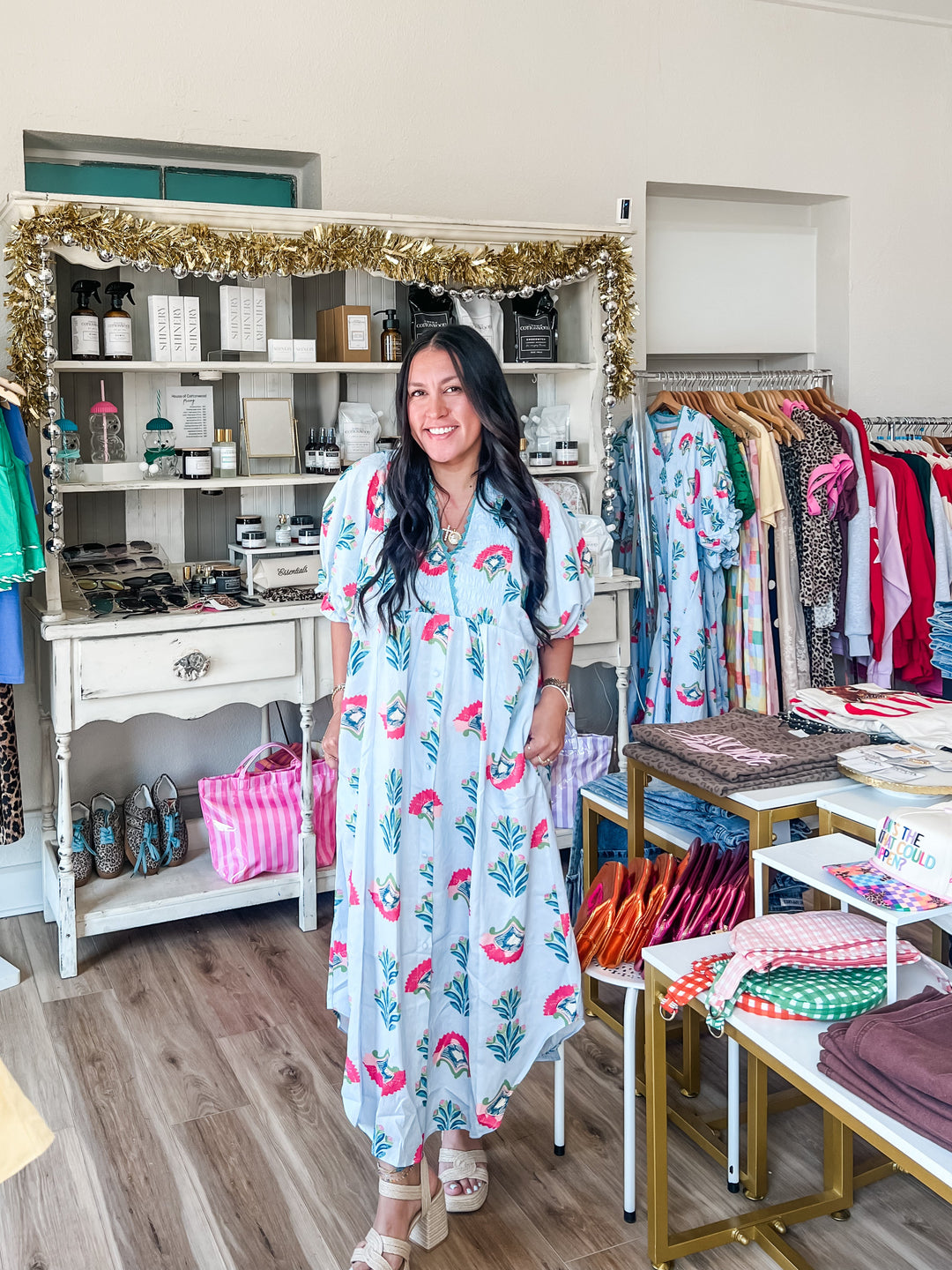 A woman stands in a boutique wearing a light blue maxi dress with a pink floral print. The shop is filled with clothing racks, shelves, and display tables.