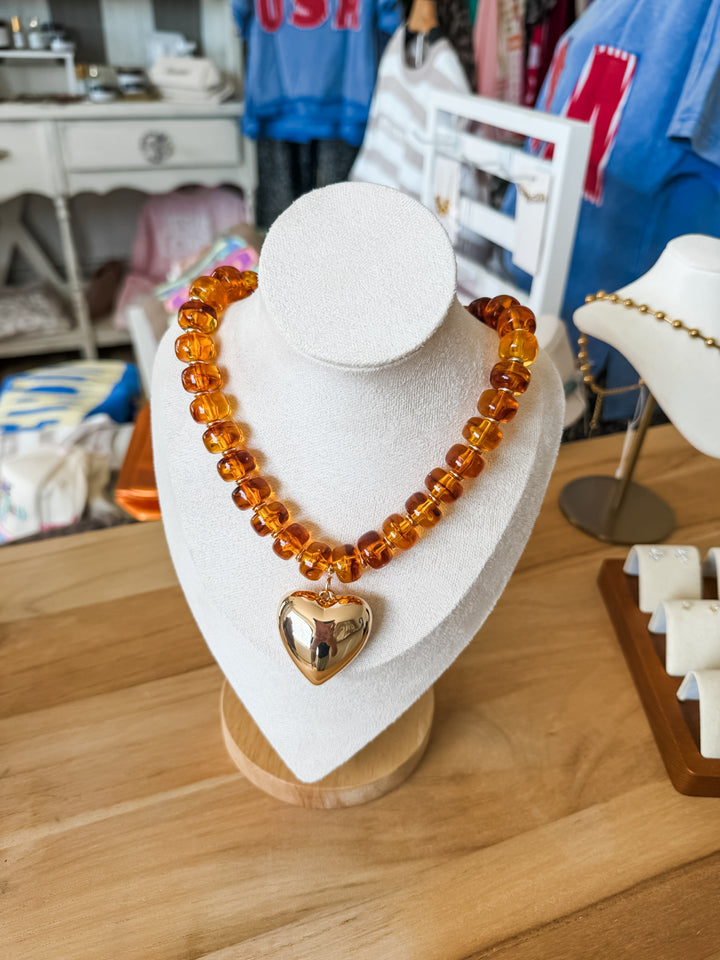 A chunky amber glass bead necklace with a polished gold heart pendant, displayed on a white jewelry bust on a wooden table.