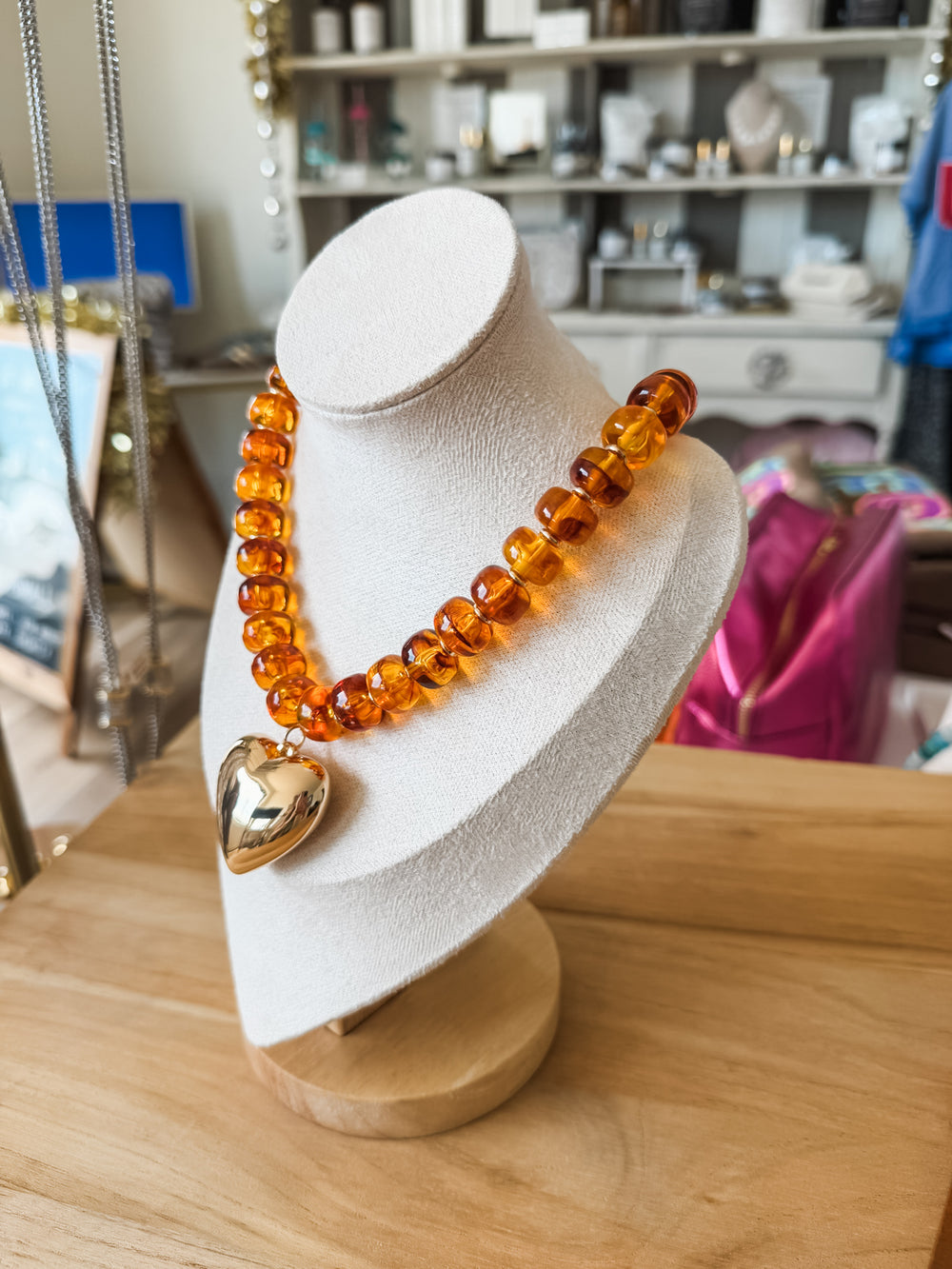 A translucent amber-colored beaded necklace with a polished gold heart pendant, displayed on a white jewelry bust on a wooden table.