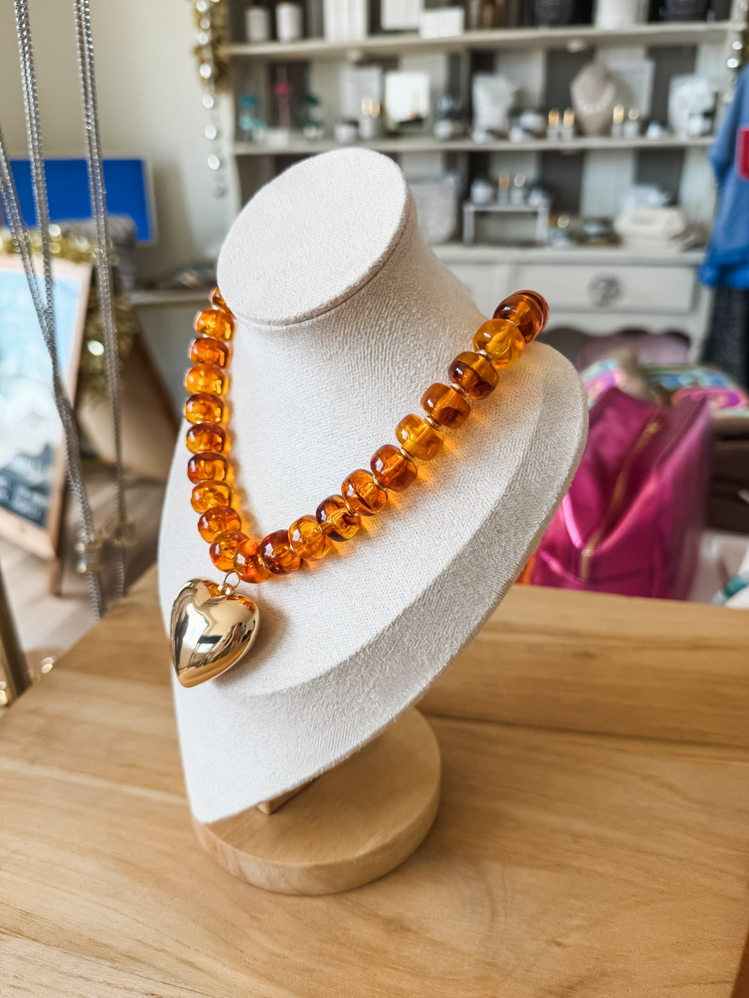 A translucent amber-colored beaded necklace with a polished gold heart pendant, displayed on a white jewelry bust on a wooden table.