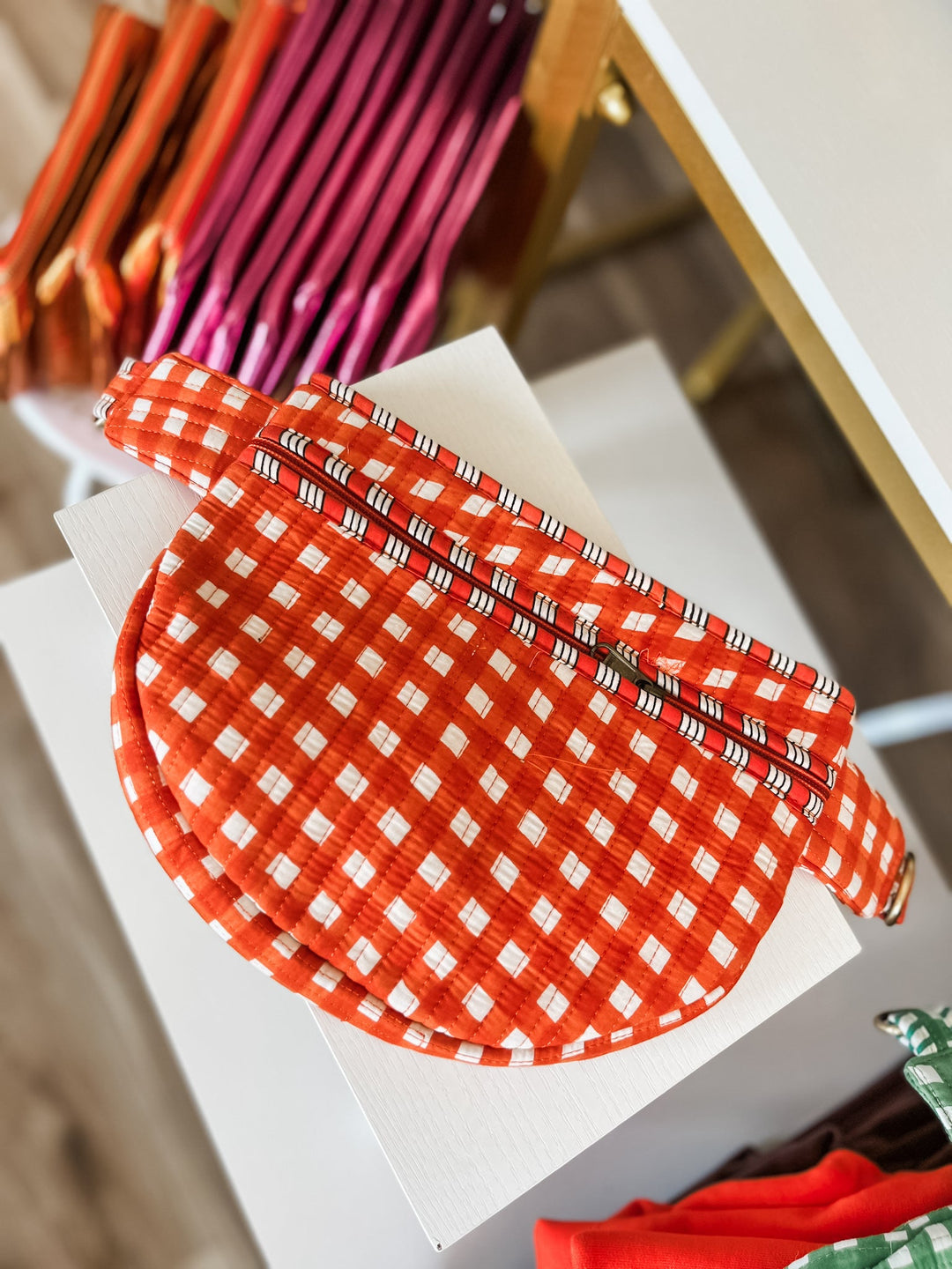 A close-up of an orange and white gingham patterned fabric item, likely a pouch or accessory, resting on a white surface with other colorful items blurred in the background.