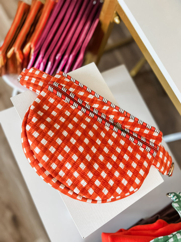 A close-up of an orange and white gingham patterned fabric item, likely a pouch or accessory, resting on a white surface with other colorful items blurred in the background.