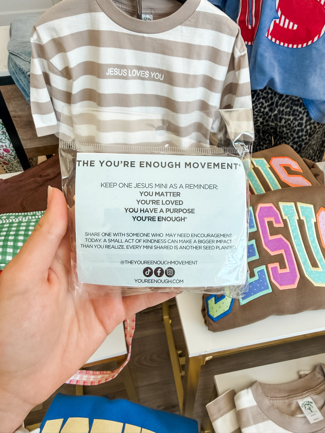 A hand holds a clear plastic bag containing a beige and white horizontally striped t-shirt with the text &