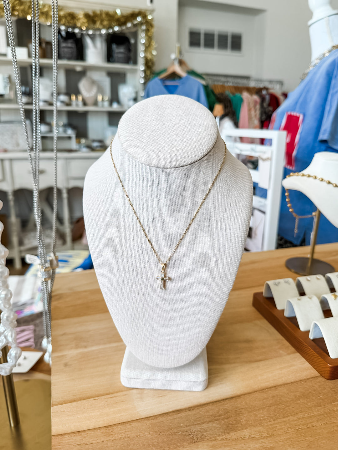 A delicate gold cross pendant necklace displayed on a white fabric jewelry bust, set on a wooden counter inside a retail store.