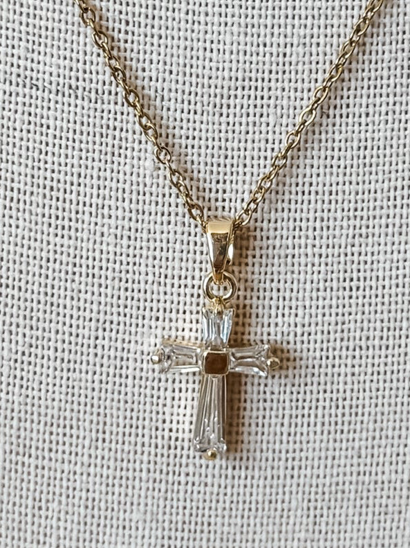 A gold cross pendant necklace with clear crystal-like accents, displayed on a textured off-white fabric background.