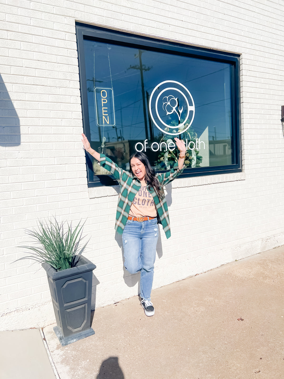 A woman posing outside a white brick building with a large window, wearing a green and white plaid flannel shirt over a tan graphic tee, blue jeans, and sneakers. The window displays the logo and name 'of one lion'.