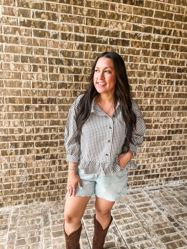 A woman wearing a black and white gingham button-down shirt, light-wash denim shorts, and brown cowboy boots.