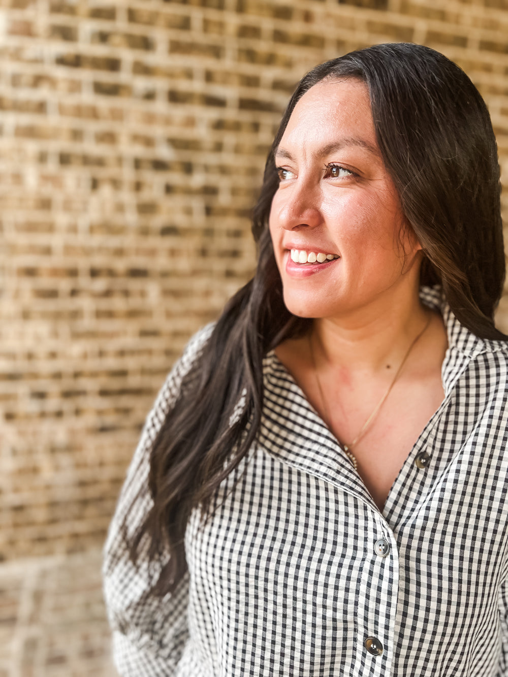 A woman with long dark hair smiling and looking to the side, wearing a black and white gingham button-up shirt, set against a blurred brick wall background.