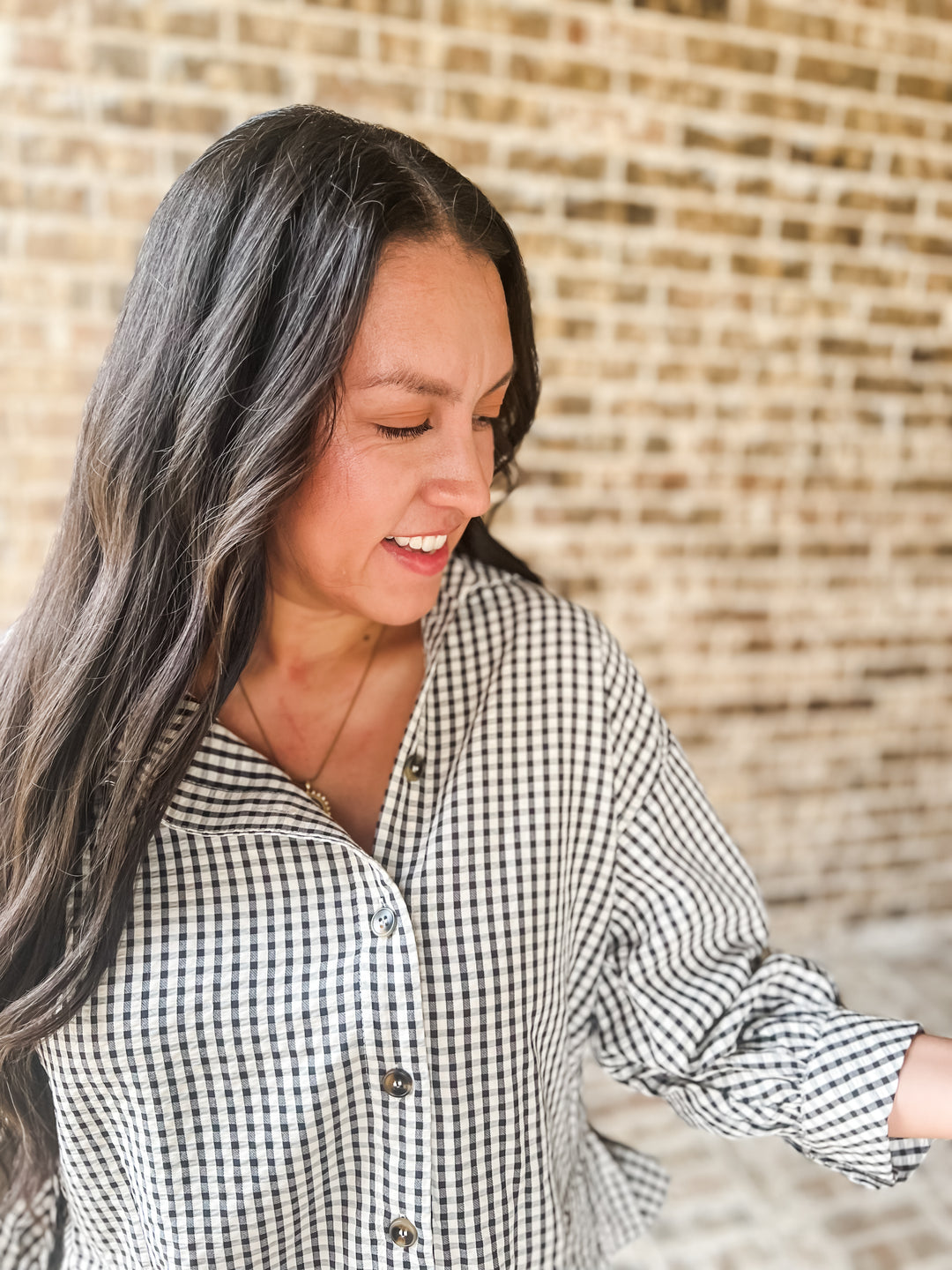 A woman with long dark hair wearing a black and white gingham button-down shirt, standing in front of a light brick wall.