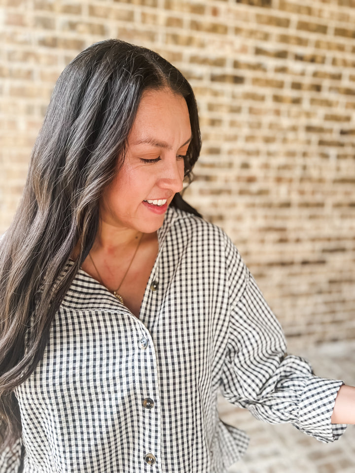 A woman with long dark hair wearing a black and white gingham button-down shirt, standing in front of a light brick wall.