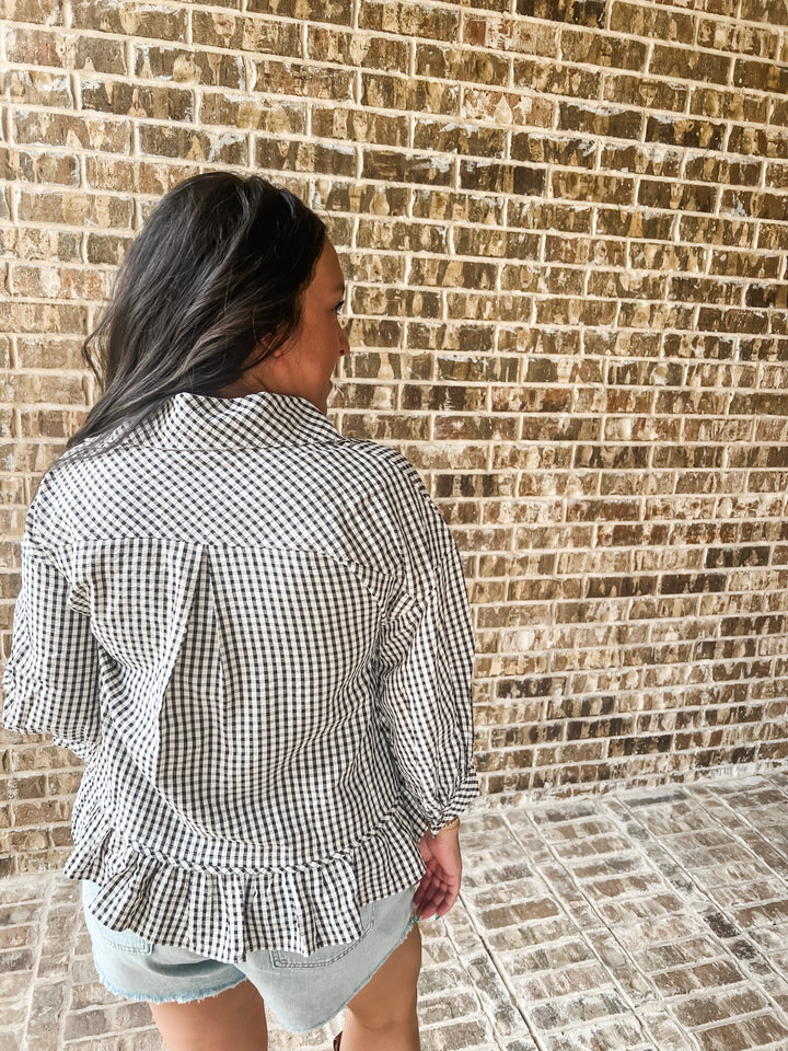 A woman viewed from behind wearing a black and white gingham check long-sleeve blouse with a ruffled hem, paired with denim shorts, standing against a brick wall.