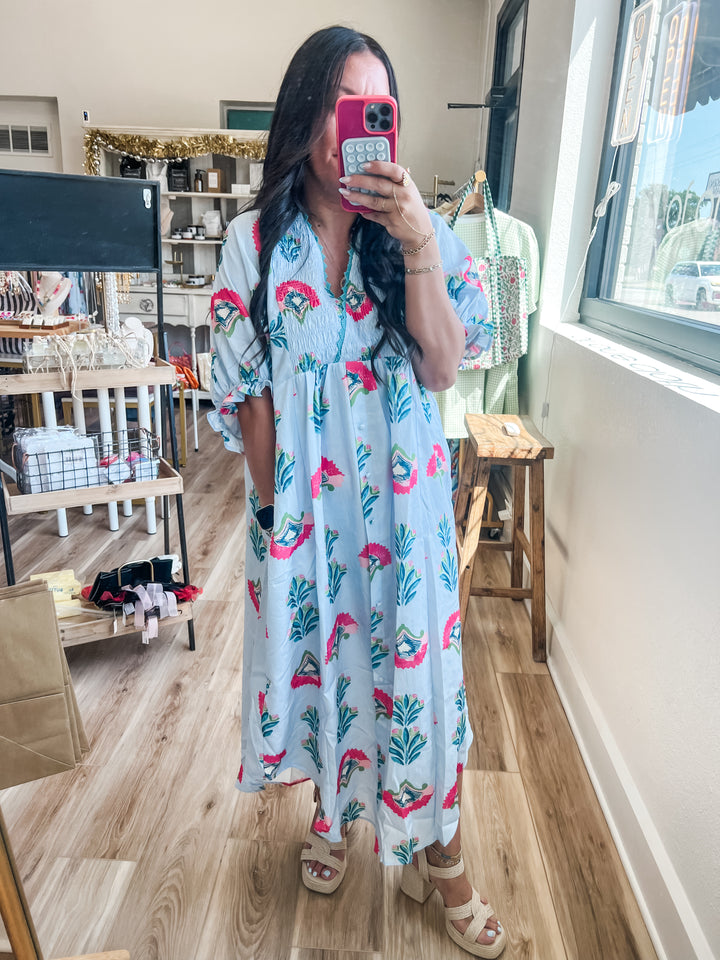 A woman takes a mirror selfie in a boutique while wearing a light blue maxi dress with a pink and green tropical print. The dress features a smocked bodice and short flutter sleeves.
