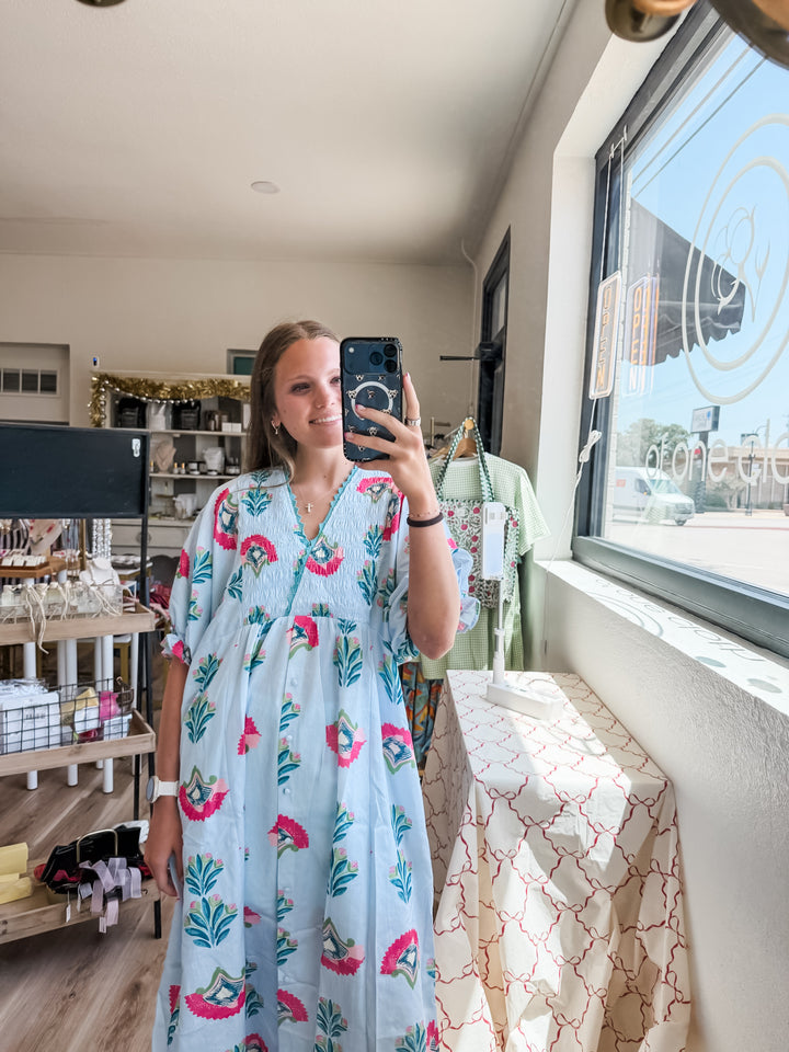 A woman takes a mirror selfie in a boutique wearing a light blue maxi dress with a pink and green tropical floral print. The shop interior is visible in the background, including shelves, clothing racks, and a large window.