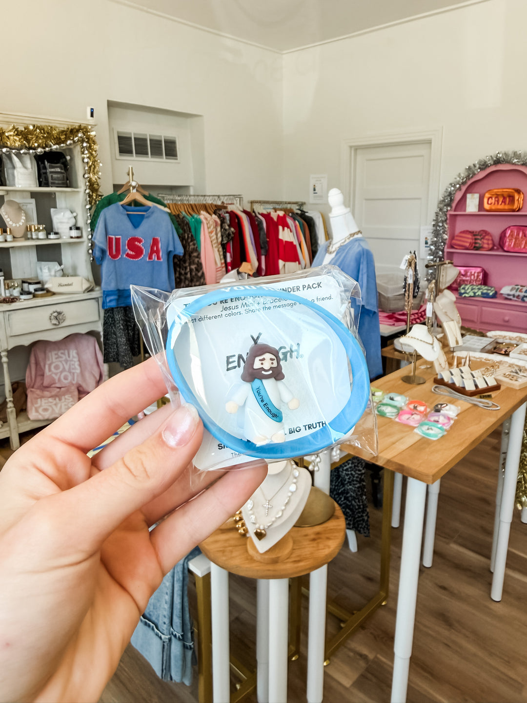 A hand holds a packaged mini Jesus and acrylic bracelet in a boutique interior. The Jesus has a light blue accent and a white clothing with a bracelet and the text 'You're Enough'.