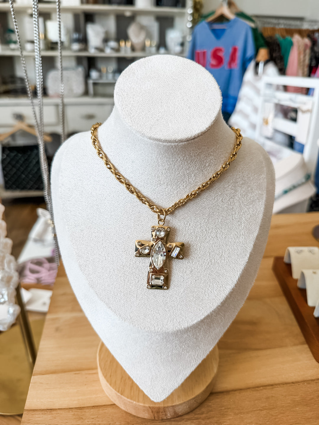 A gold-tone cross pendant necklace with a clear marquise-cut center stone, displayed on a white jewelry bust in a retail store setting.
