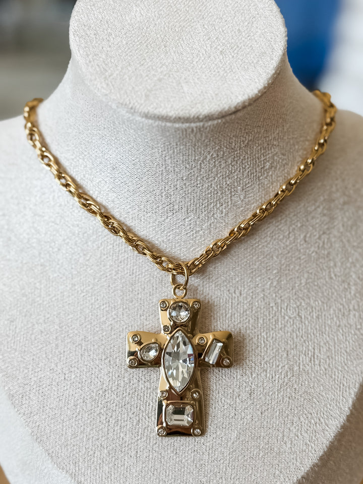 A gold cross pendant necklace with clear marquise and round crystals, displayed on a textured white jewelry bust.