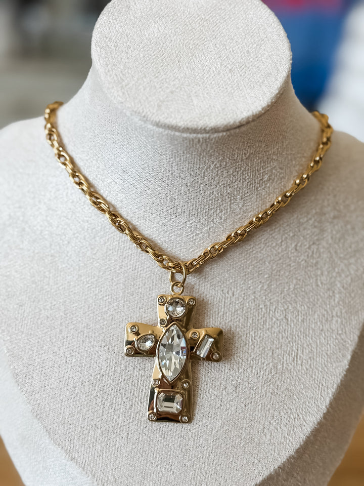 A gold cross pendant necklace with clear rhinestones, displayed on a cream-colored fabric jewelry bust.