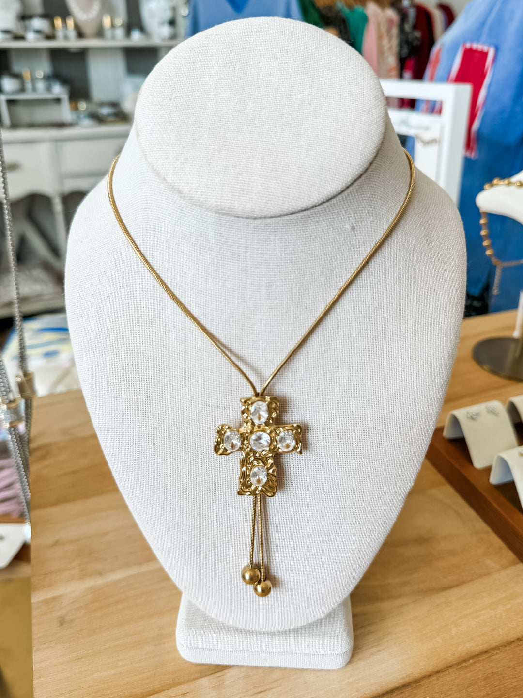 A gold cross pendant necklace with clear rhinestones and dangling gold beads, displayed on a white fabric jewelry bust in a retail setting.