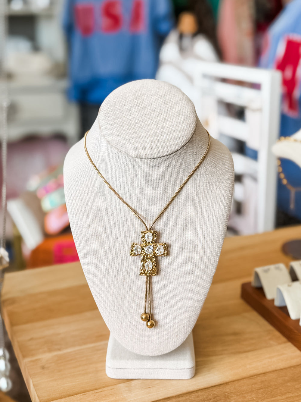 A gold cross pendant necklace with clear rhinestones and dangling bead accents, displayed on a white jewelry bust on a wooden table.