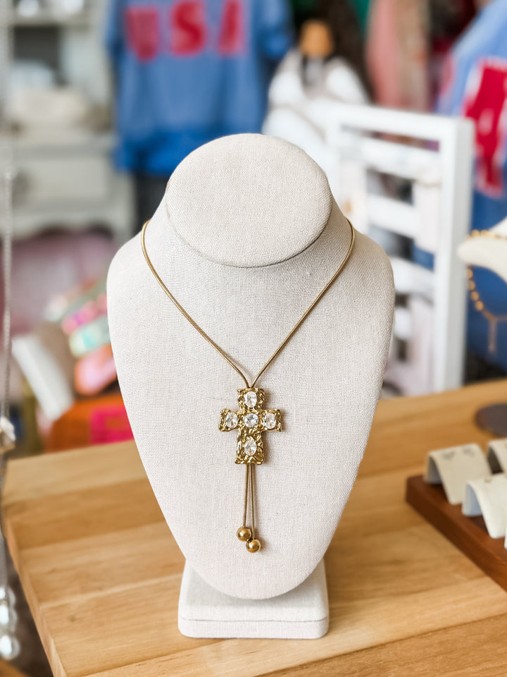 A gold cross pendant necklace with clear rhinestones and dangling bead accents, displayed on a white jewelry bust on a wooden table.