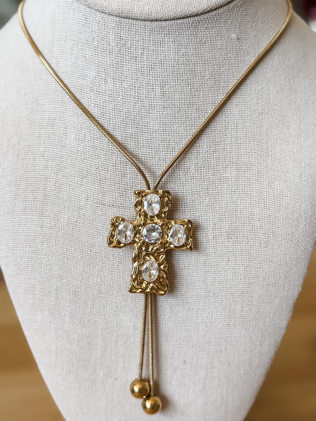 Gold cross pendant necklace with clear rhinestones and dangling gold beads, displayed on a cream fabric jewelry bust.