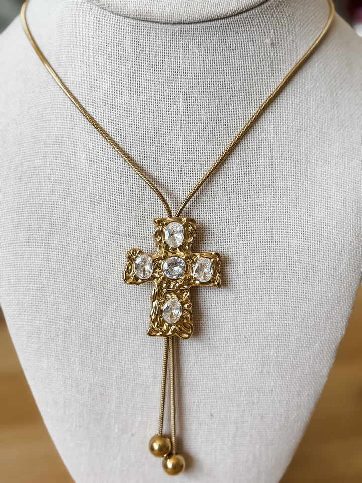 Gold cross pendant necklace with clear rhinestones and dangling gold beads, displayed on a cream fabric jewelry bust.