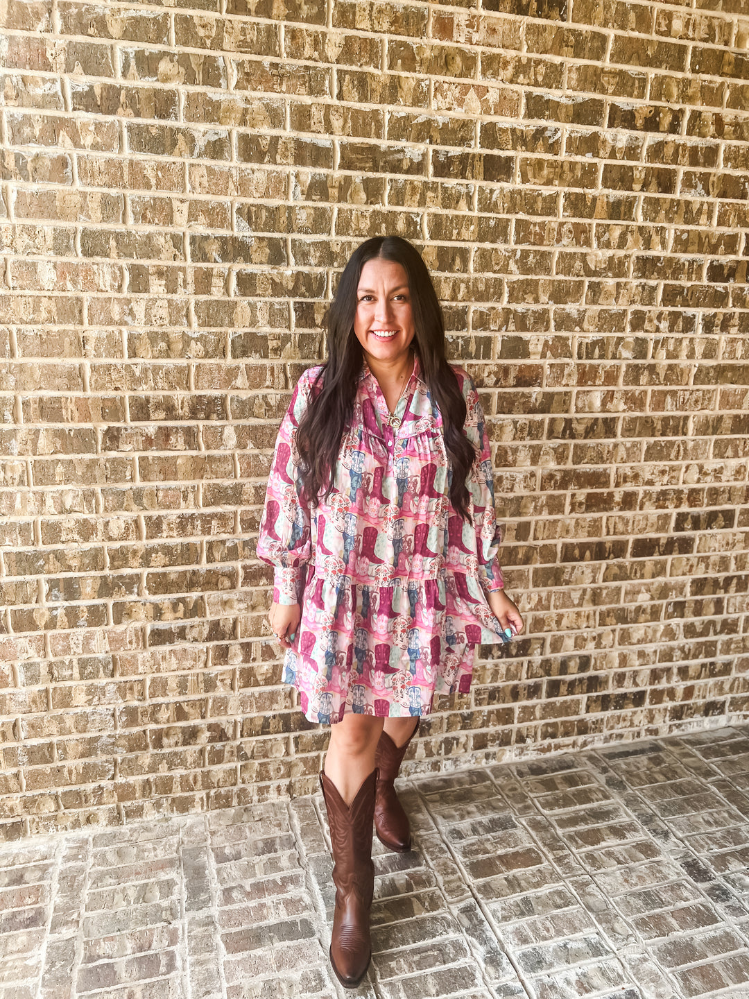 A woman stands in front of a tan brick wall wearing a pink and blue boot patterned long-sleeve mini dress with brown cowboy boots.
