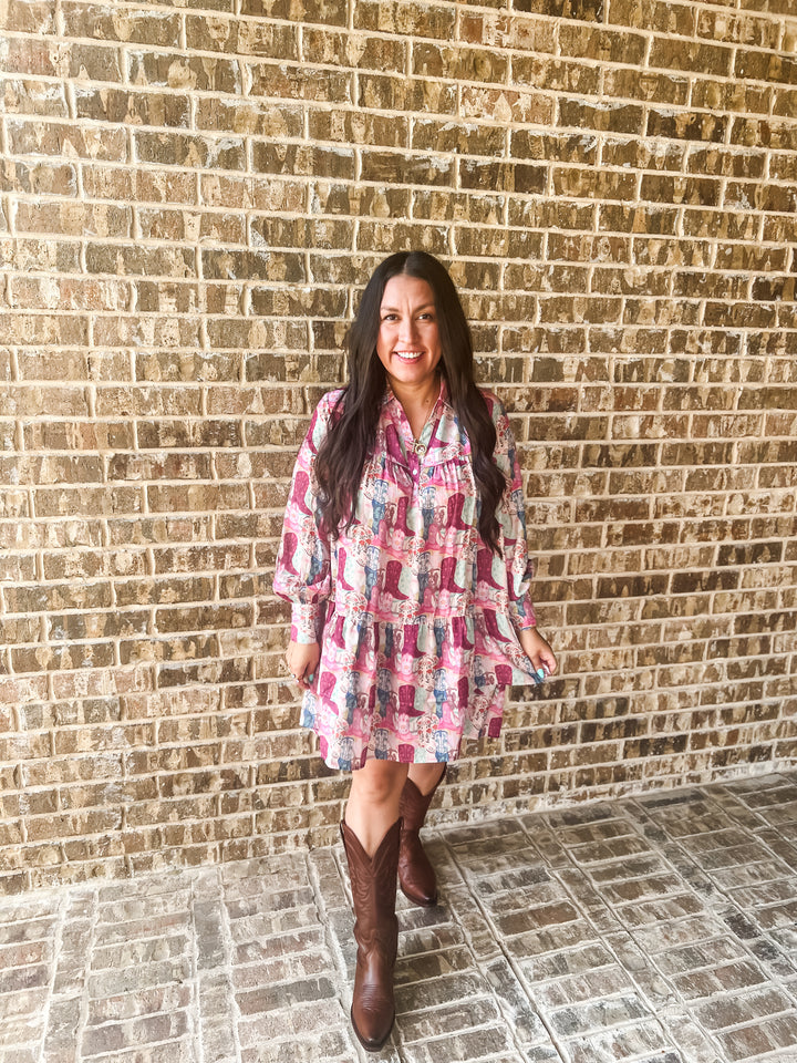 A woman stands in front of a tan brick wall wearing a pink and blue boot patterned long-sleeve mini dress with brown cowboy boots.