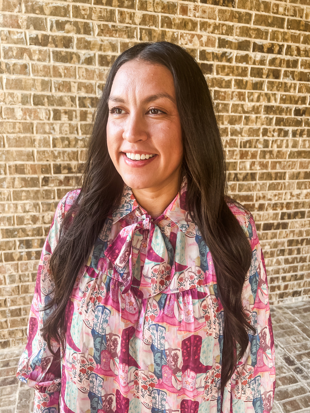 A smiling woman with long dark hair wearing a pink and blue patterned dress with a tie-neck detail, standing in front of a tan brick wall.