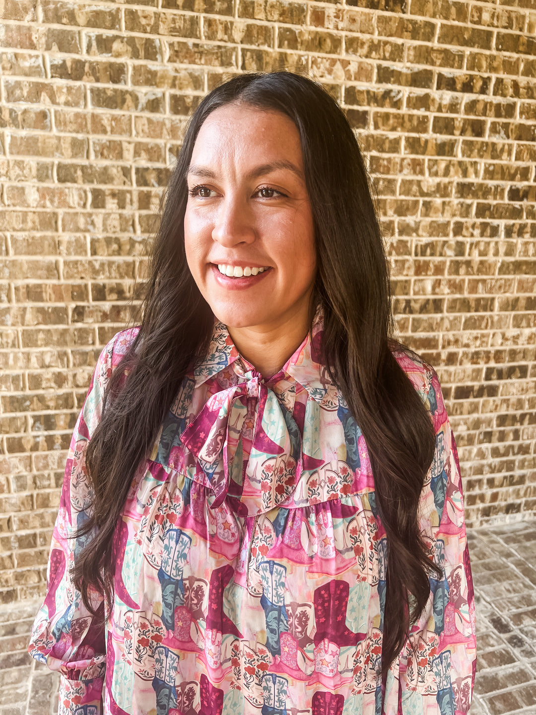 A smiling woman with long dark hair wearing a pink and blue patterned dress with a tie-neck detail, standing in front of a tan brick wall.