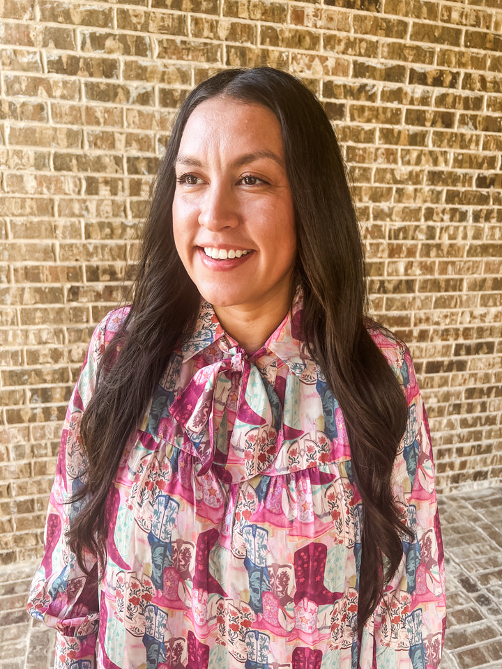 A smiling woman with long dark hair wearing a pink and blue patterned dress with a tie-neck detail, standing in front of a tan brick wall.