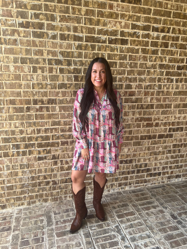 A woman stands in front of a tan brick wall wearing a pink and blue floral long-sleeve mini dress with brown cowboy boots.