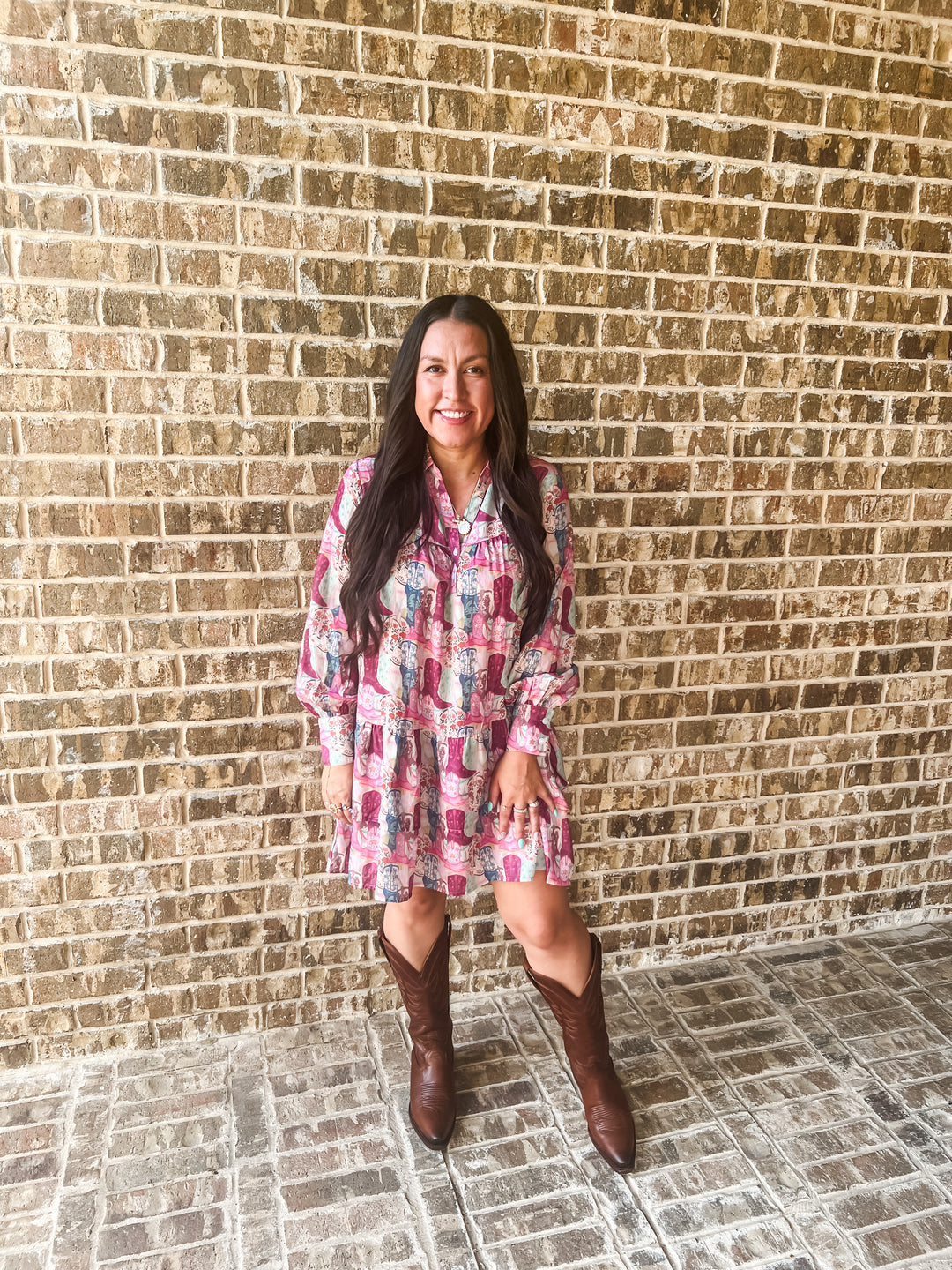 A woman stands against a tan brick wall wearing a pink and multicolor patterned long-sleeve mini dress with brown cowboy boots.