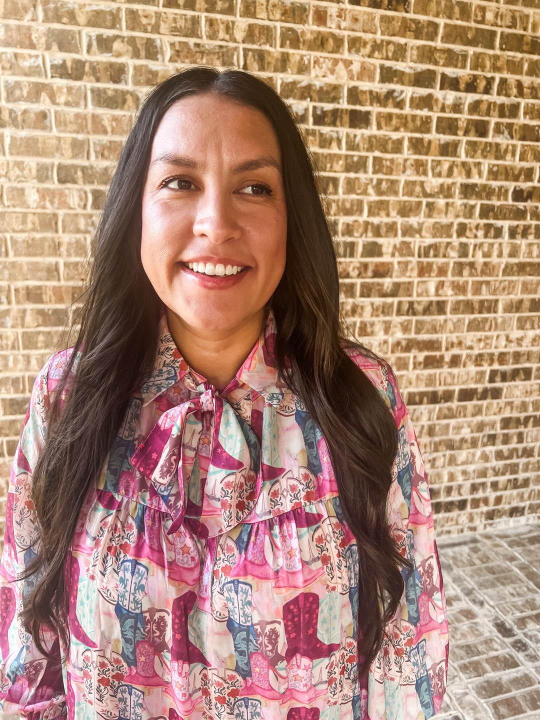 A smiling woman with long dark hair wearing a colorful pink, purple, and blue patterned boot blouse with a ruffled tie-neck collar, standing in front of a tan brick wall.