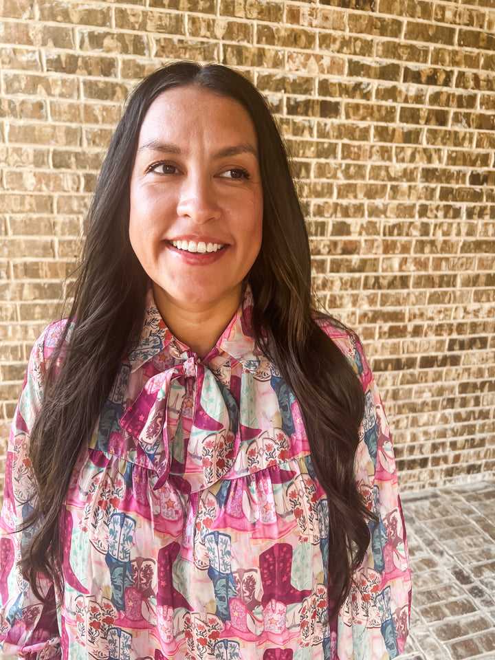A smiling woman with long dark hair wearing a colorful pink, purple, and blue patterned boot blouse with a ruffled tie-neck collar, standing in front of a tan brick wall.
