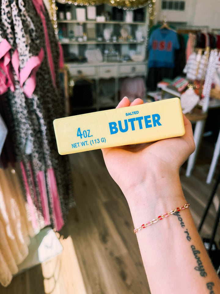 A hand holding a 4 oz stick of salted butter squishy in a retail store setting with clothing racks in the background.
