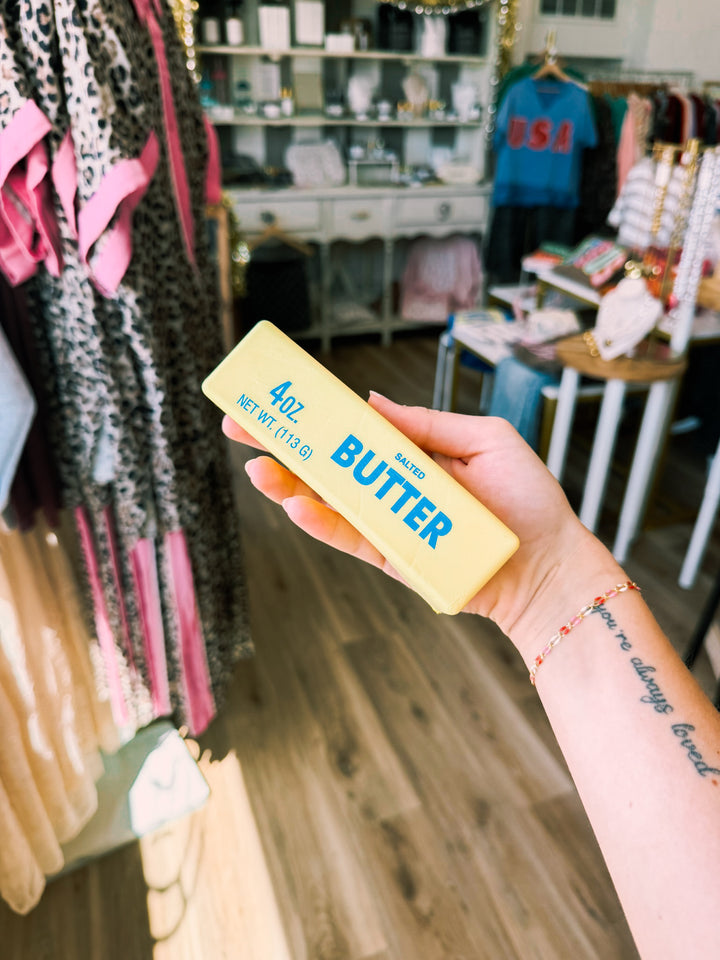 A hand holding a 4 oz stick of Butter brand salted butter squishy, with a boutique clothing store visible in the blurred background.