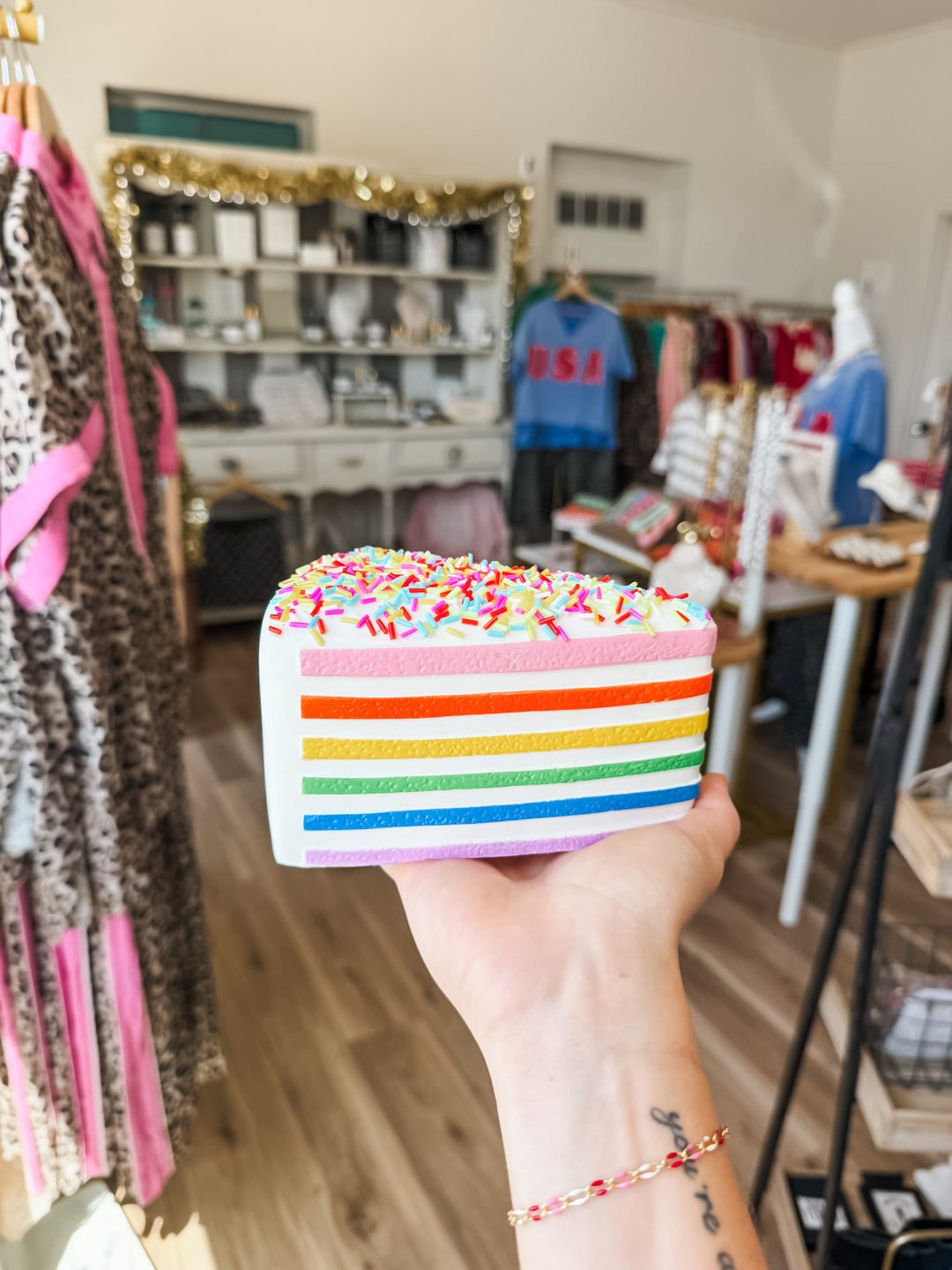 A hand holds a triangular cake-shaped squishy with rainbow-colored horizontal stripes and a white top covered in multicolored sprinkles, set against a blurred boutique interior.