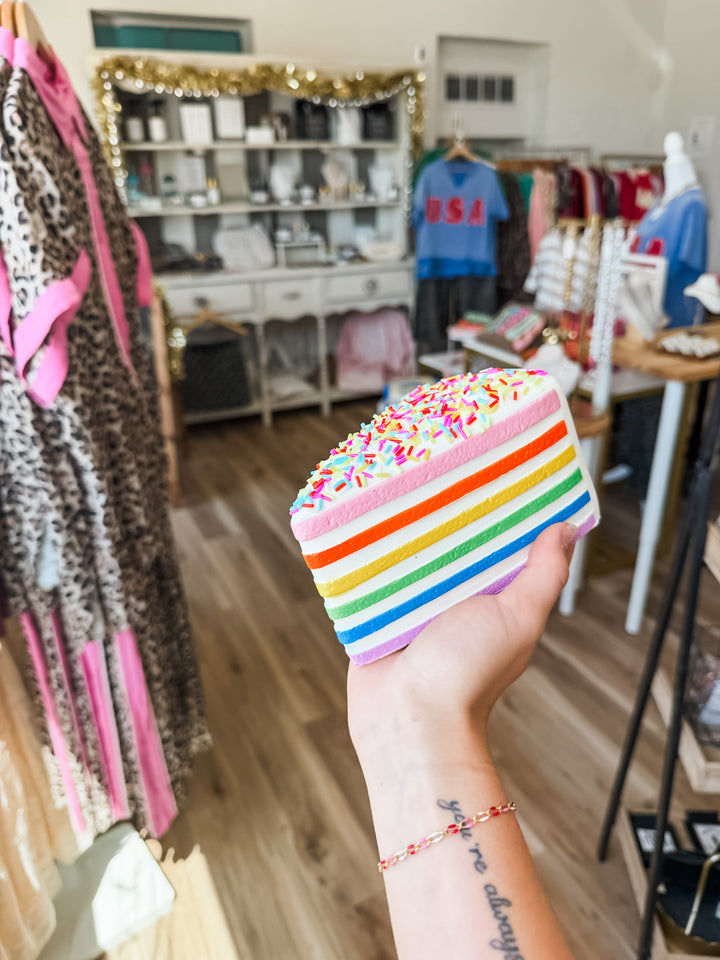 A hand holds a small rainbow-striped cake slice toy squishy with colorful sprinkles in a retail store setting with clothing racks and shelves in the background.