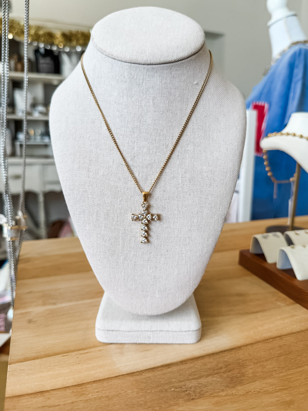 A gold cross pendant necklace adorned with clear heart shaped crystals, displayed on a white fabric jewelry bust. The bust is set on a light wooden table, with a softly blurred boutique background showing other jewelry items.
