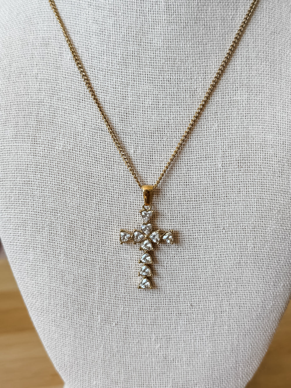 A gold cross pendant necklace adorned with clear heart shaped crystals, displayed on a textured beige fabric jewelry bust.