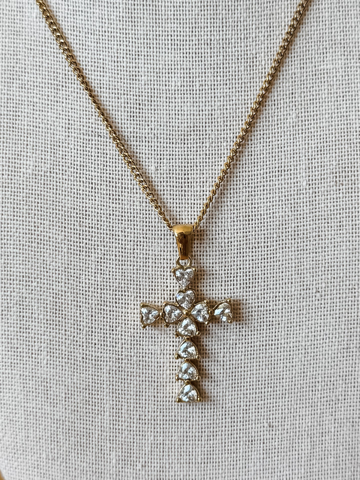 A gold cross pendant necklace adorned with clear heart-shaped crystals, displayed on a textured beige fabric background.