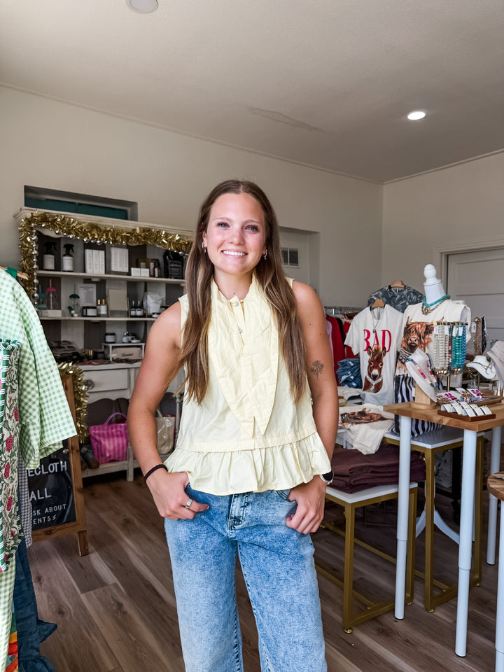 A woman with long brown hair stands smiling inside a boutique, wearing a pale yellow ruffled sleeveless blouse and light-wash blue jeans. The shop interior has wooden floors, display racks, and shelves with various items.
